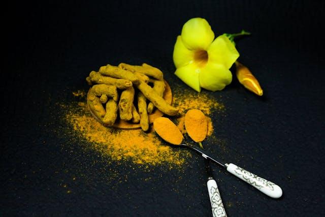 racines de curcuma et curcumine en poudre avec deux petites cuillères et une fleur jaune sur fond noir