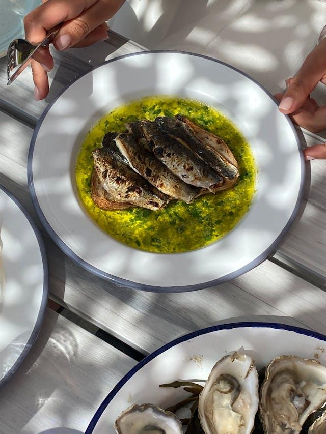 Sardines posées sur sauce jaune et verte dans une assiette blanche avec assiette d'huîtres au premier-plan