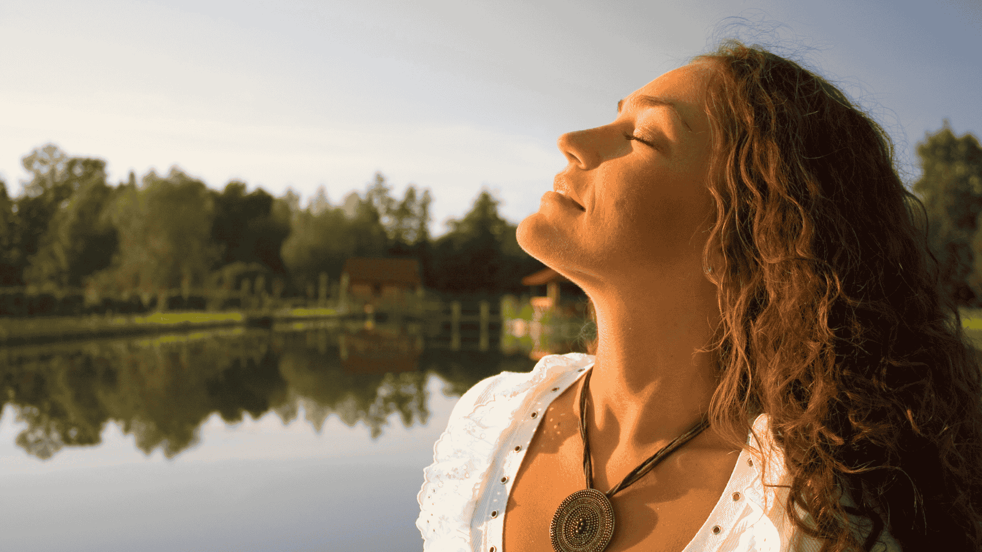 Jeune femme cheveux longs bruns ondulés visage tourné vers le soleil avec en fond de l'eau et des arbres