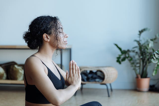 Femme noire haut noir qui médite de profil dans un salon avec plante verte au fond