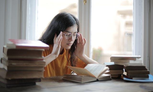 Jeune femme brune avec lunettes et tee-shirt orange qui ferme les yeux avec les mains sur ses tempes devant un bureau avec un livre ouvert et des livres fermés