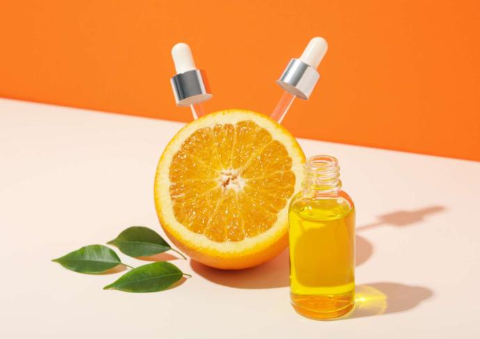 Quartier d'orange avec deux pipettes de flacon posés dessus et trois feuilles vertes et une flacon d'huile à côté sur fond orange