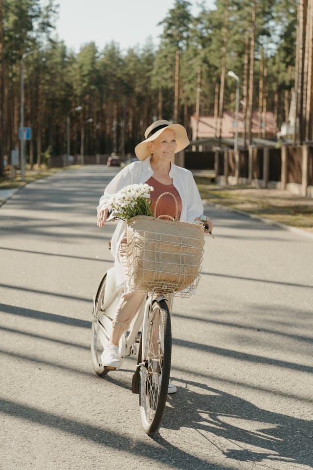Femme senior avec chapeau et panier avec bouquet de fleurs blanche qui pédale dans la rue