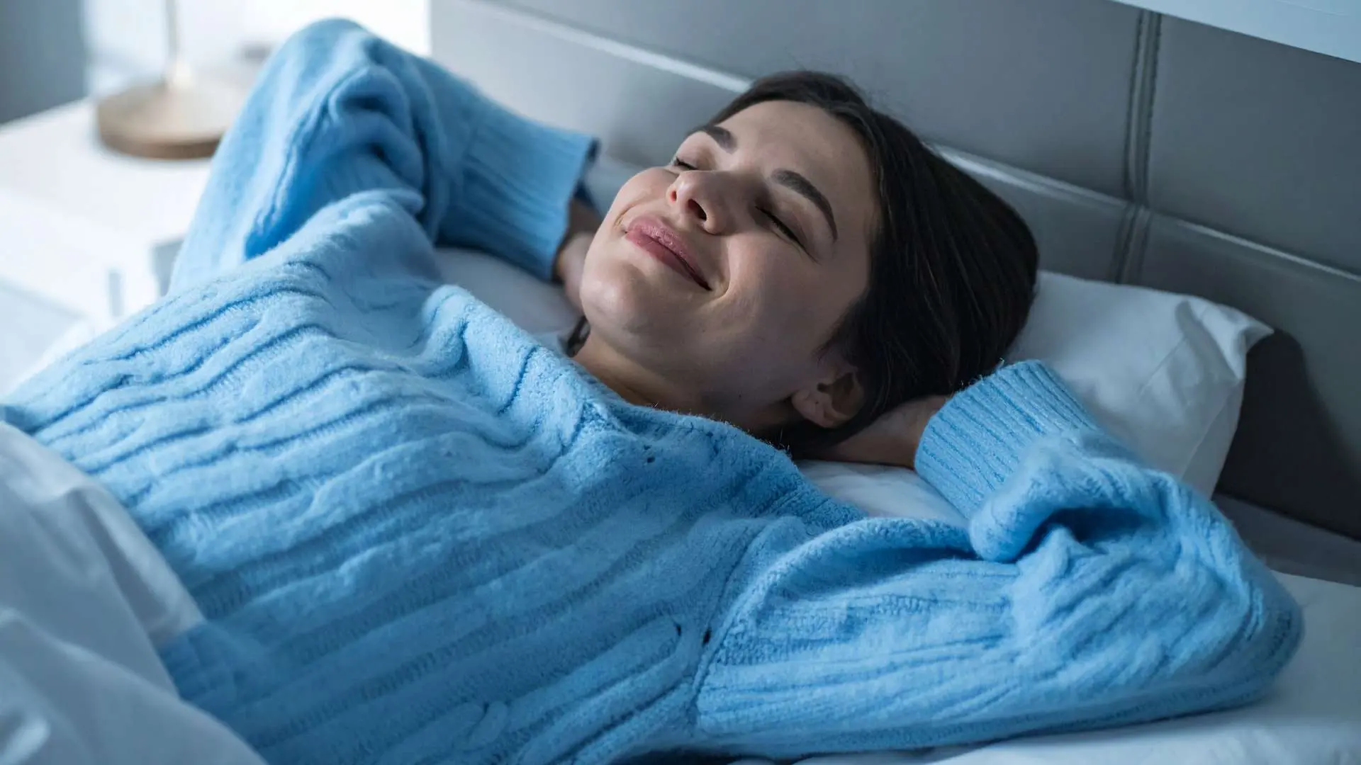 Femme  avec un pull à torsades bleu clair qui sourit les yeux fermés allongée dans son lit les mains derrière la tête