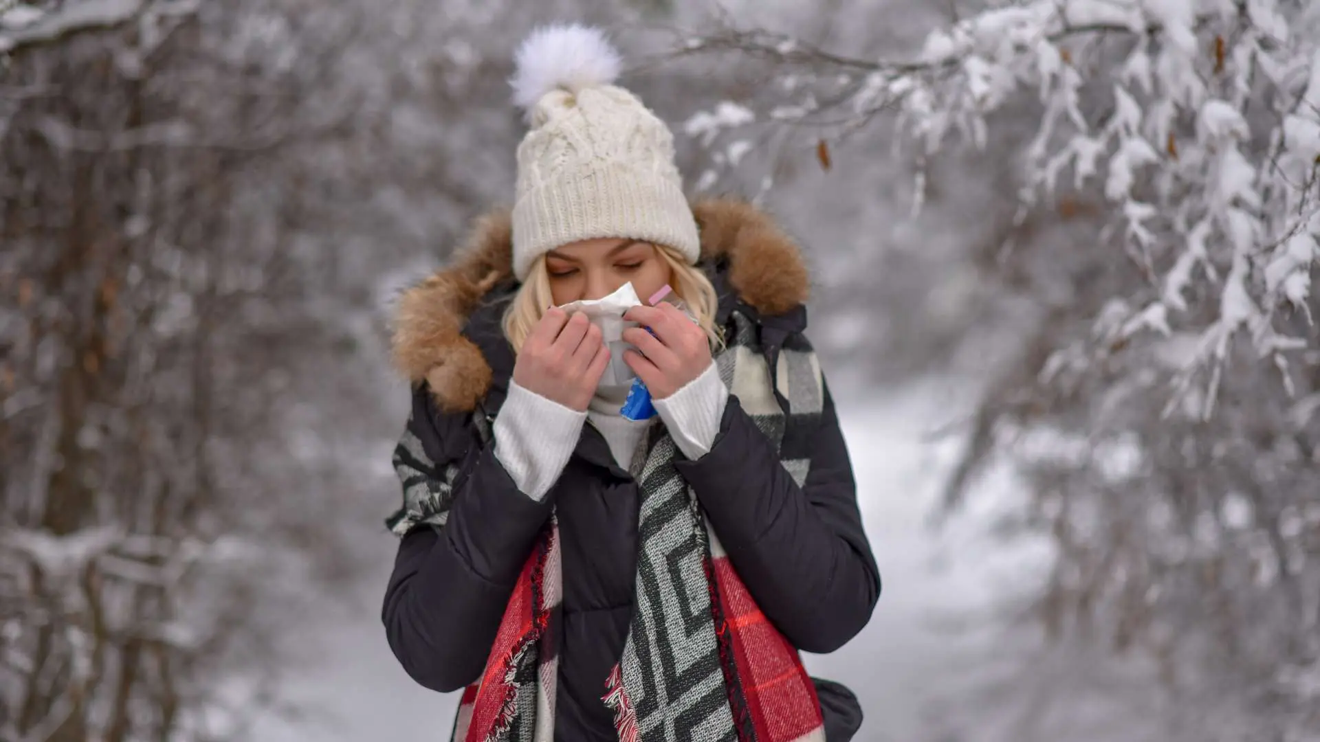 Femme avec bonnet blanc, doudoune et écharpe qui se mouche entourée d'arbres recouverts de neige